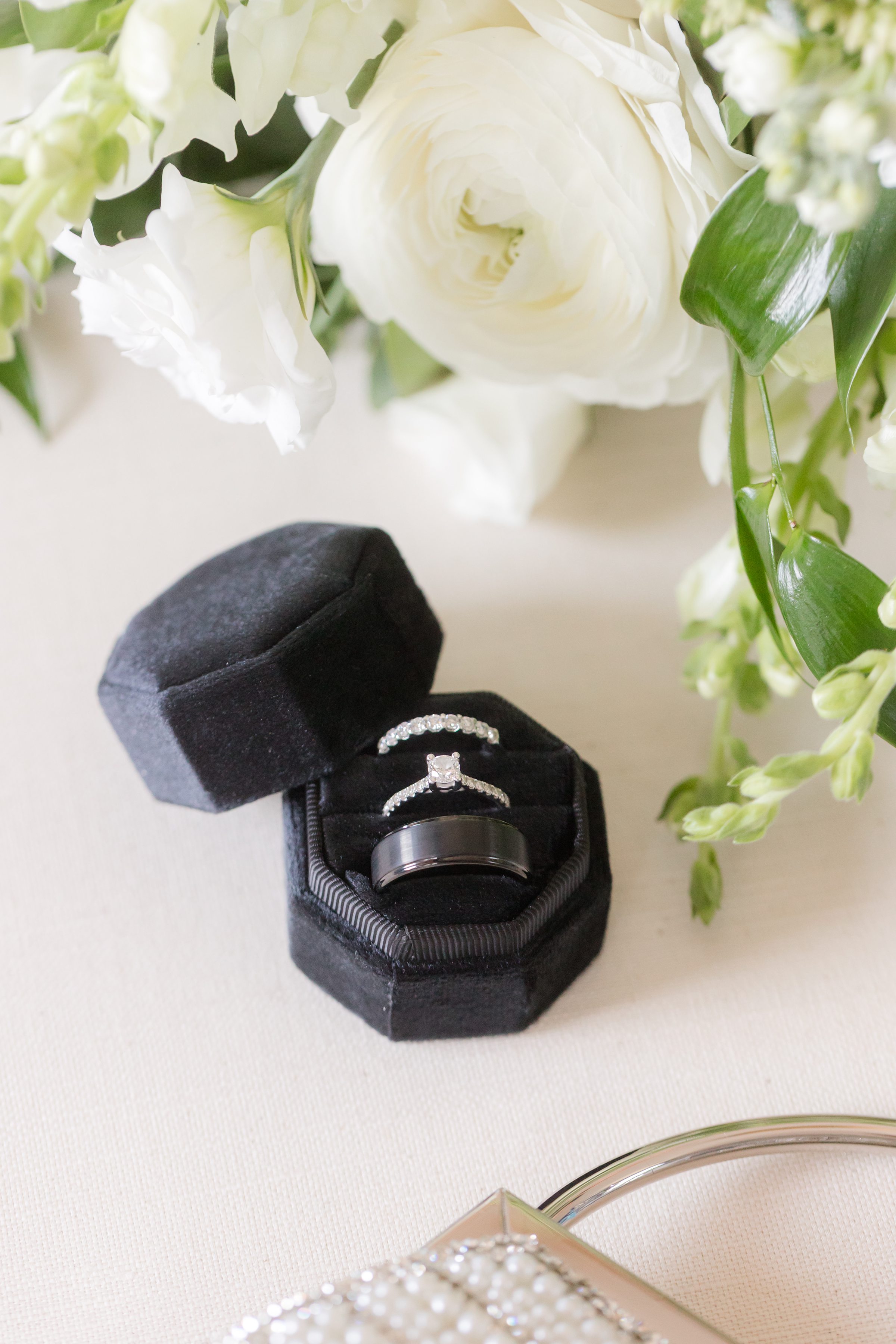 Wedding rings nestled in a black velvet box beneath soft white blooms and fresh greenery, symbolizing an elegant and timeless union.”