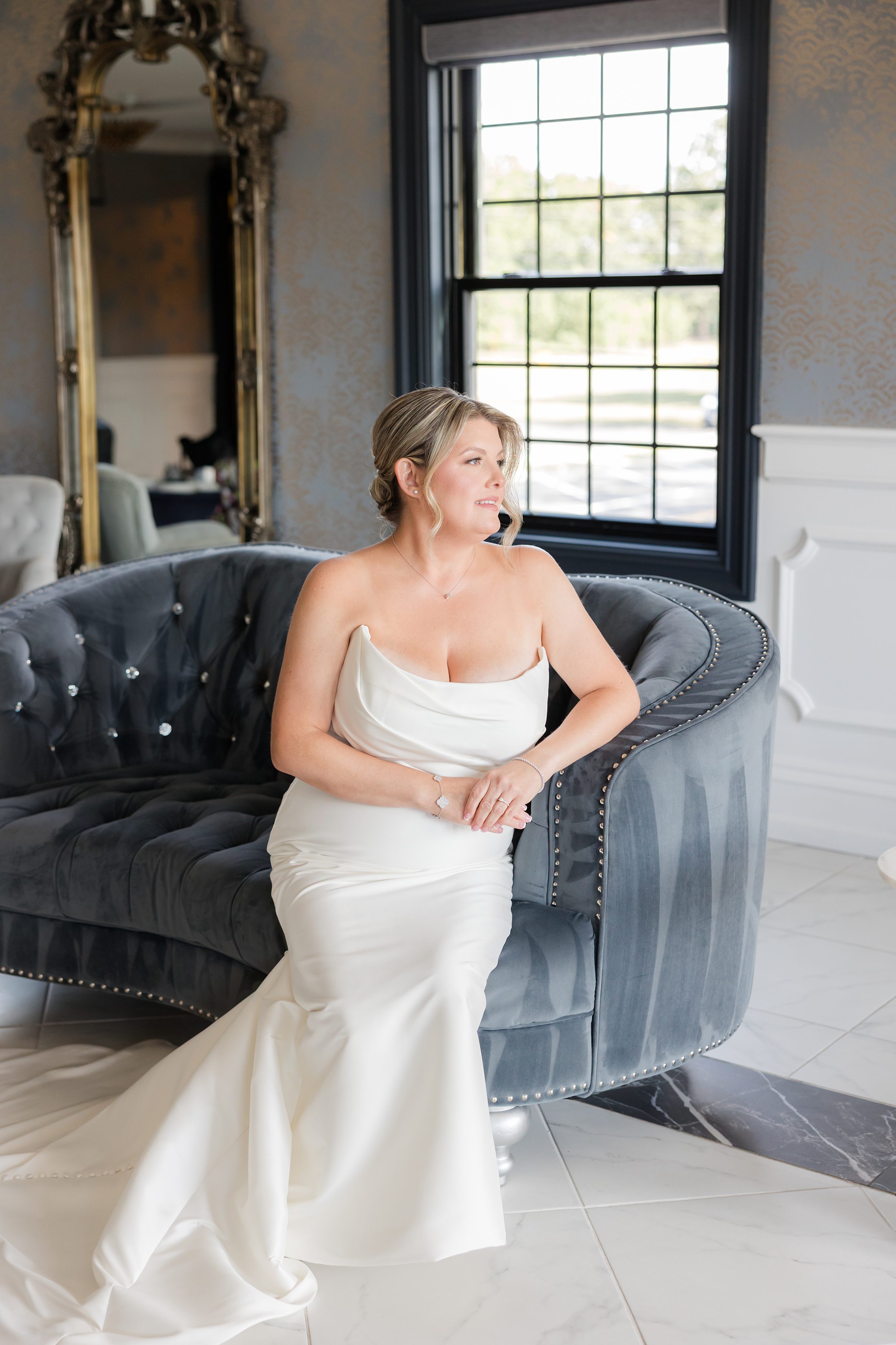 Elegant bride in a strapless ivory gown seated on a plush velvet sofa, softly gazing toward the light, surrounded by refined interiors and natural window light—capturing a quiet, romantic moment before the ceremony