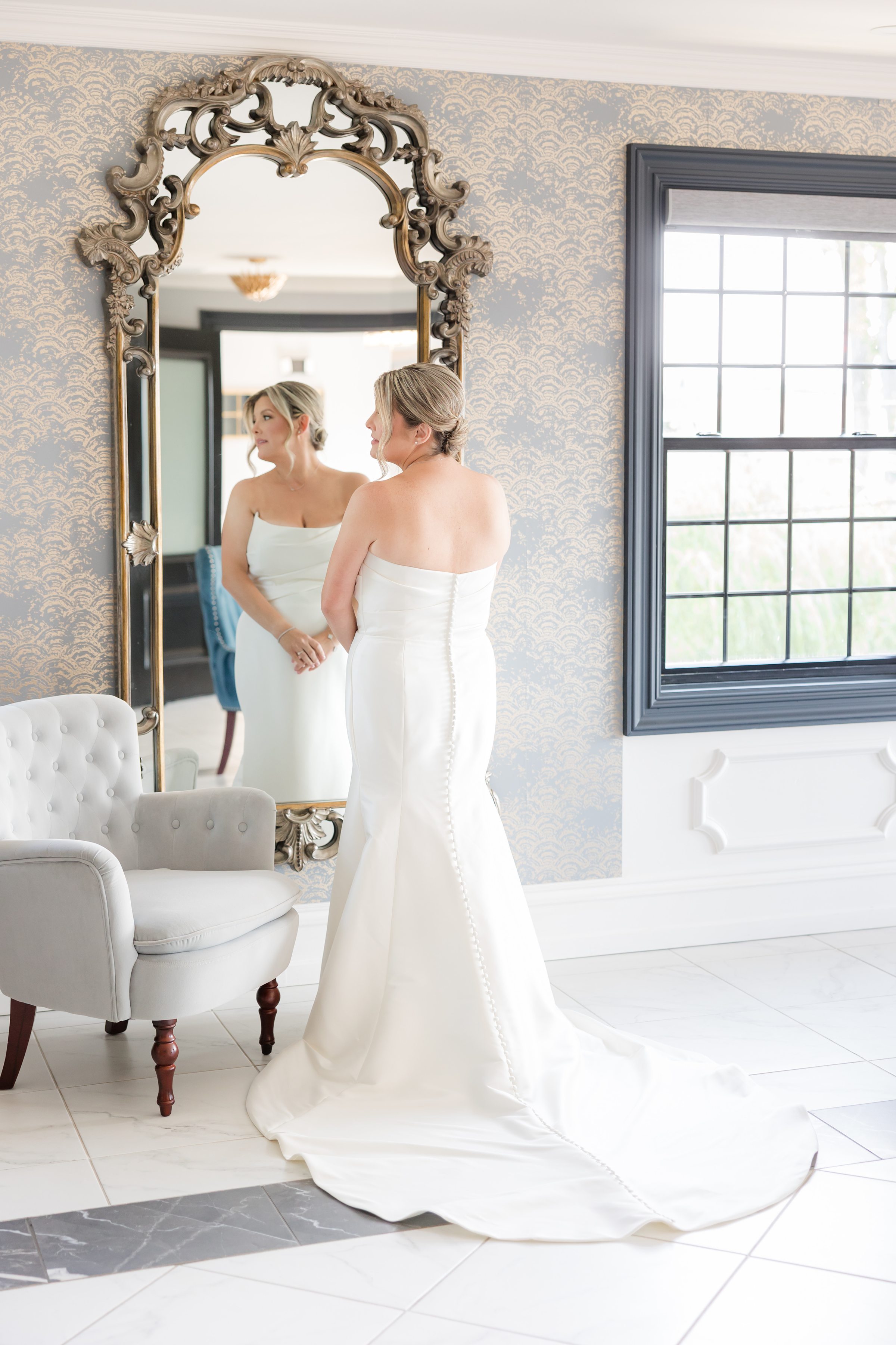 Bride stands gracefully before an ornate mirror, her reflection capturing the quiet anticipation and elegance of her wedding day