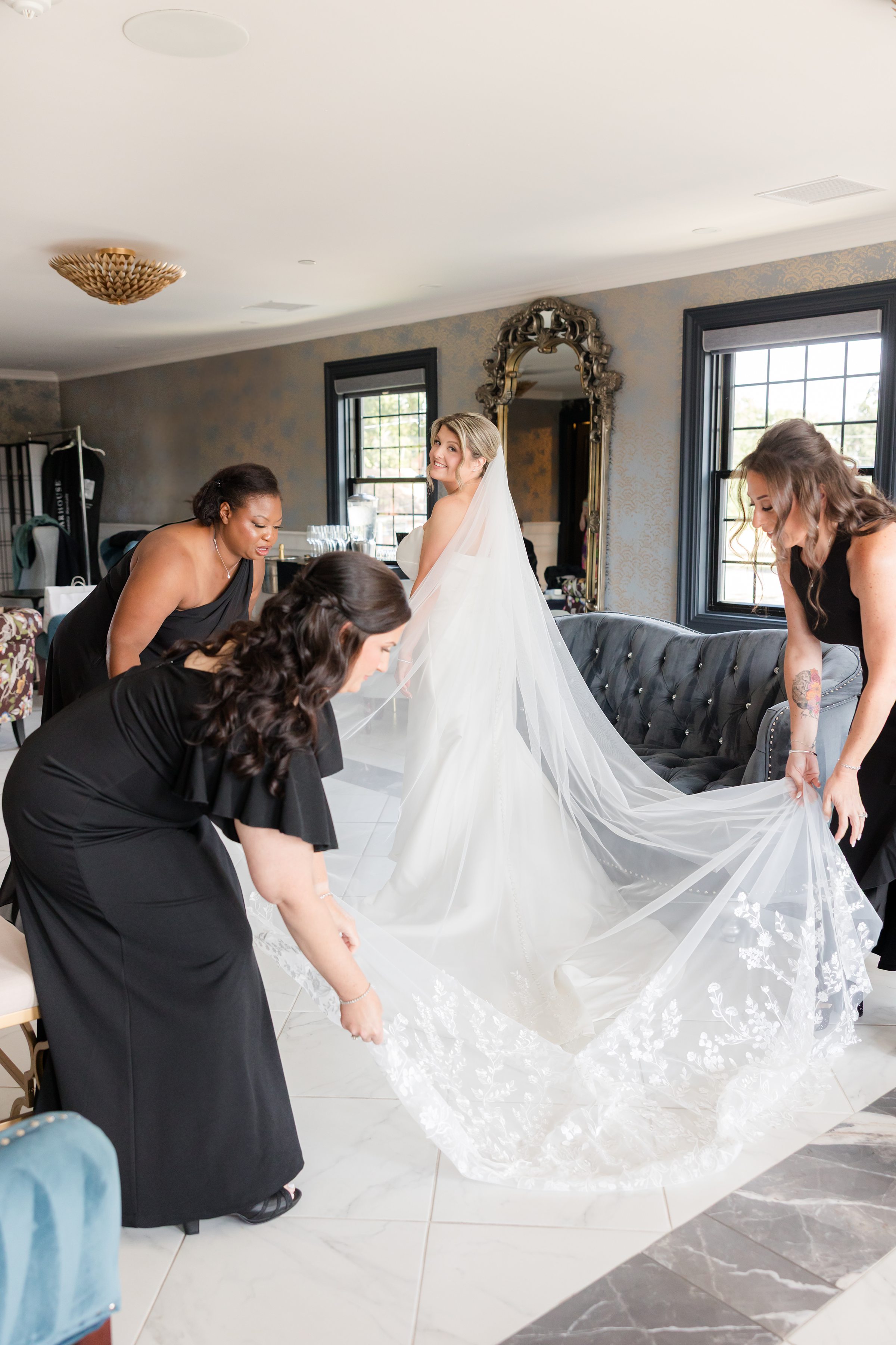 Bride stands gracefully in her gown as her bridesmaids lovingly adjust her flowing veil, sharing a quiet, intimate moment before she walks down the aisle