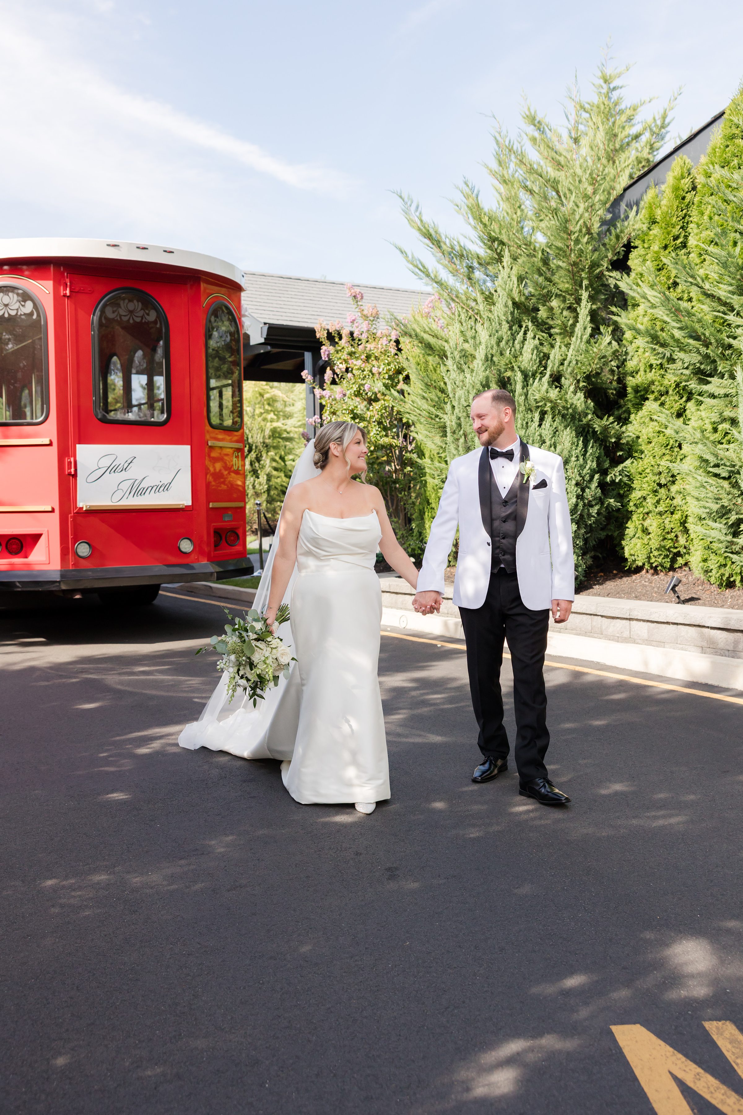 Bride and groom walk hand in hand down a sunlit path beside a red “Just Married” trolley, smiling at each other as they begin their journey together in love