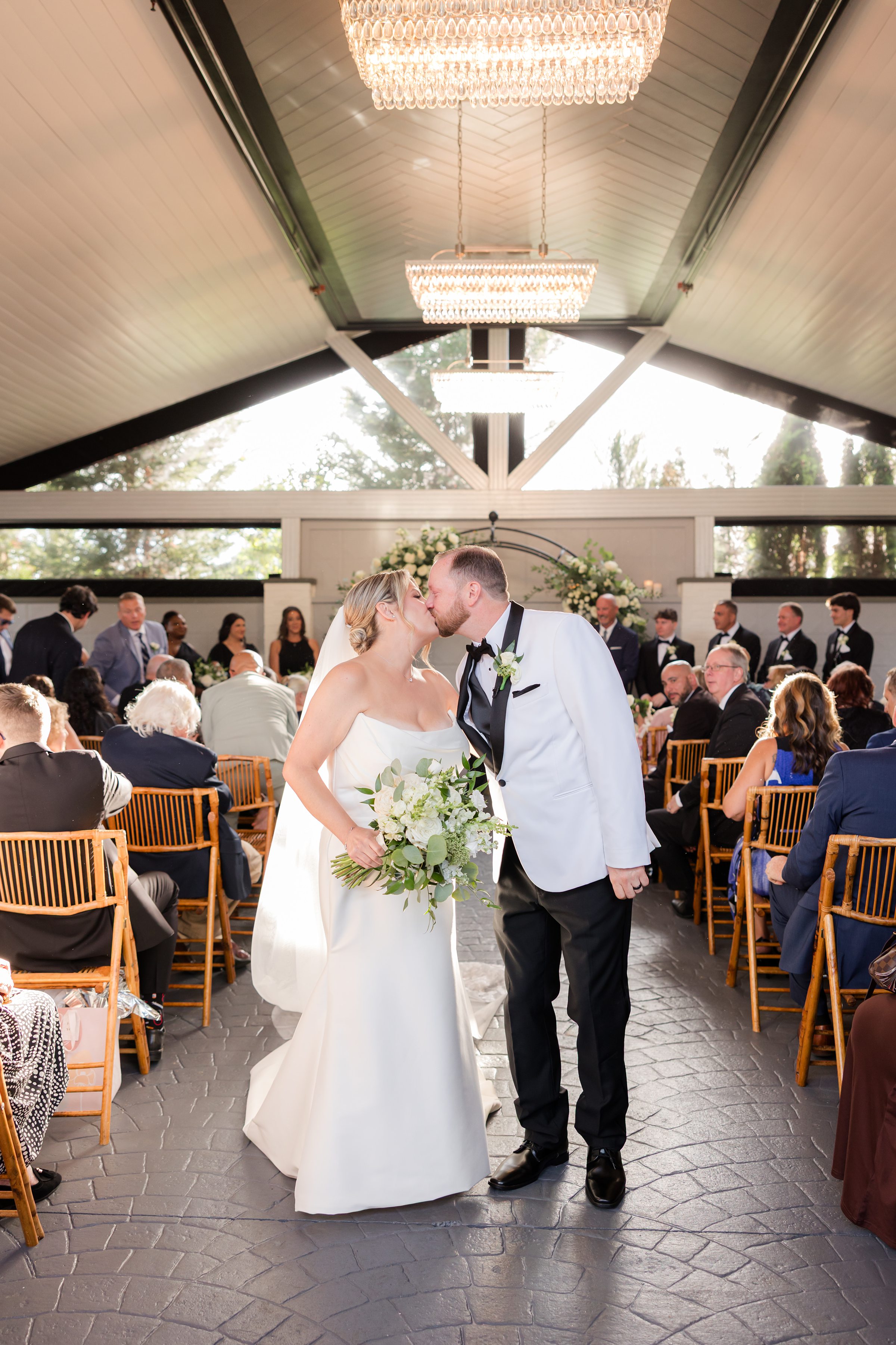 Bride and groom share a tender kiss beneath glowing chandeliers, surrounded by loved ones as sunlight pours into their wedding ceremony