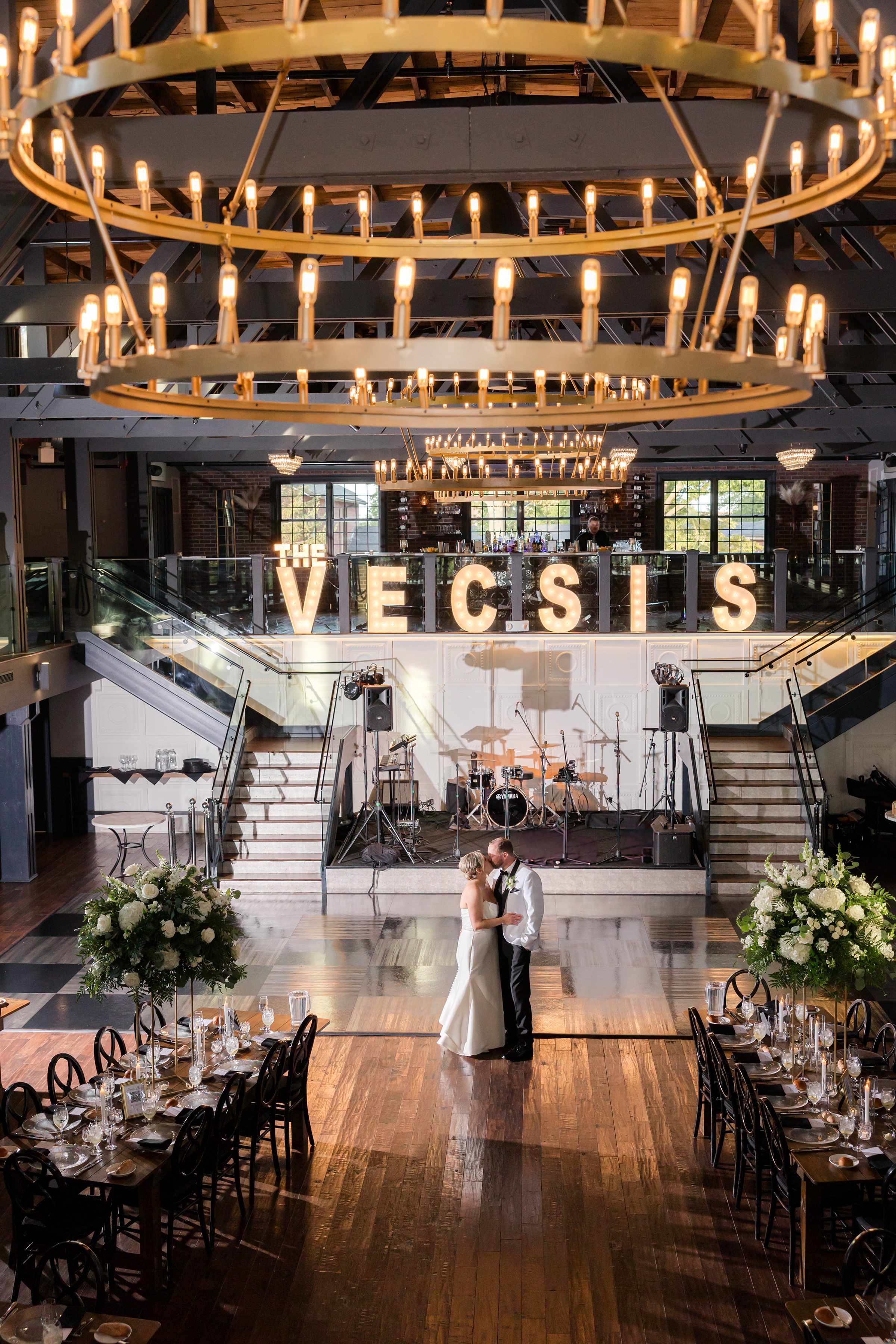 Beneath glowing chandeliers, the bride and groom share a tender kiss at the center of their reception, surrounded by warm light and love