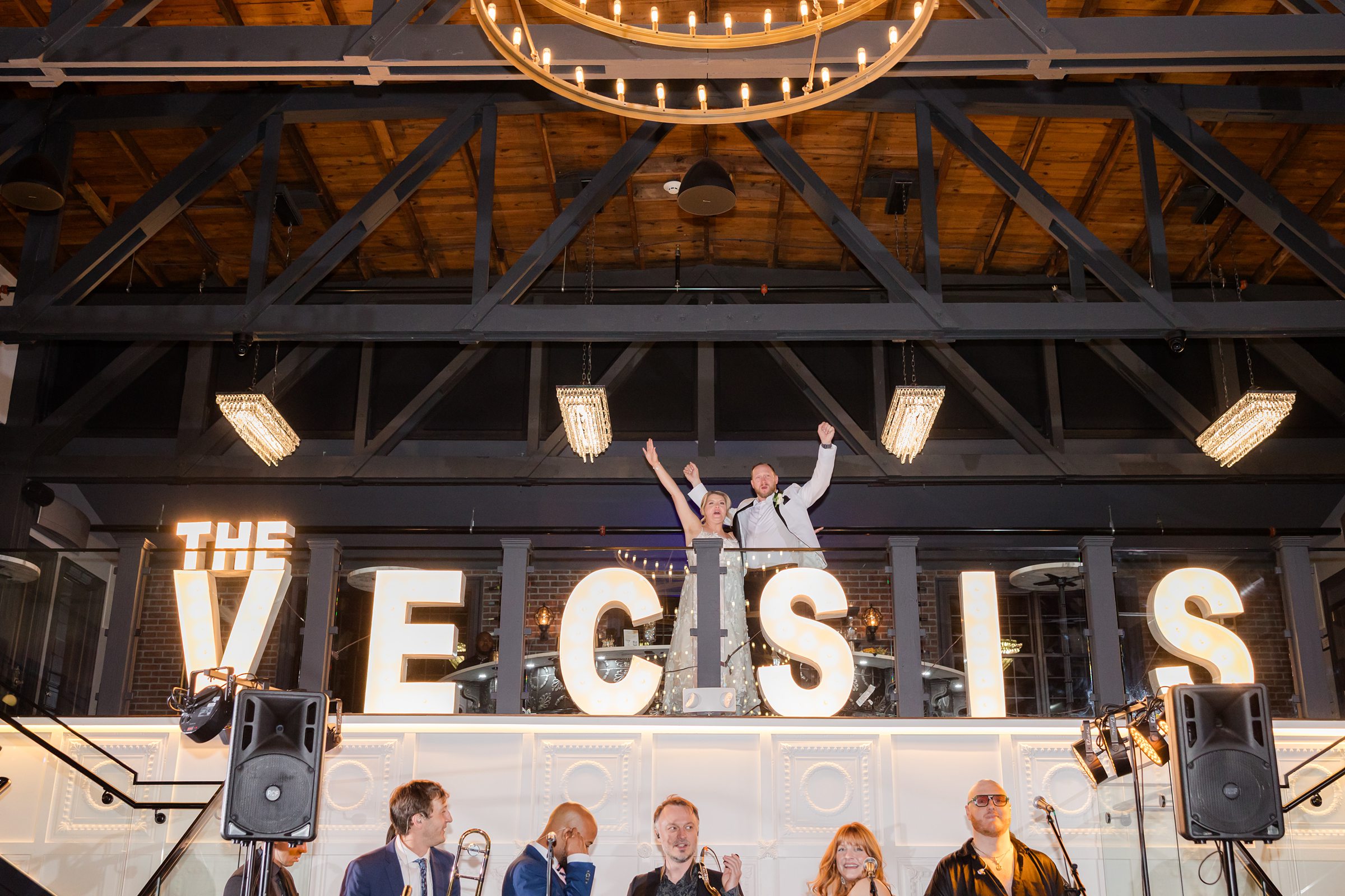 Bride and groom raise their arms in joy above glowing and celebrating their love as music fills the room below.