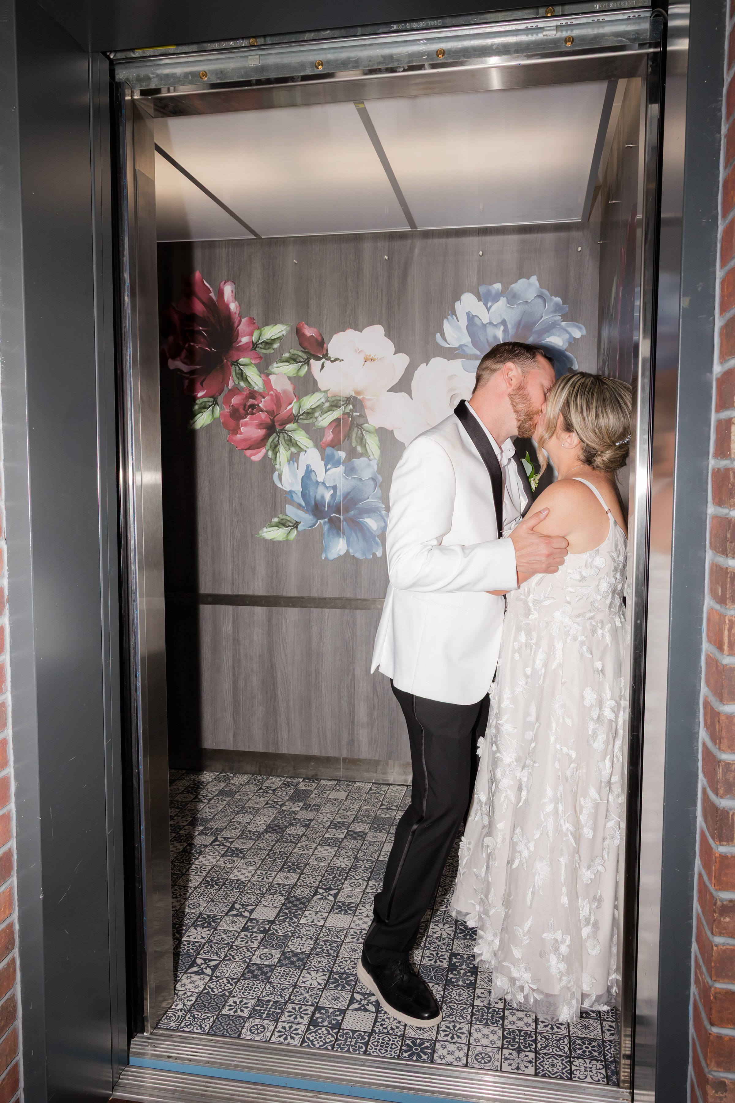 Newlyweds steal an intimate kiss in a quiet elevator, wrapped in each other against a soft floral backdrop
