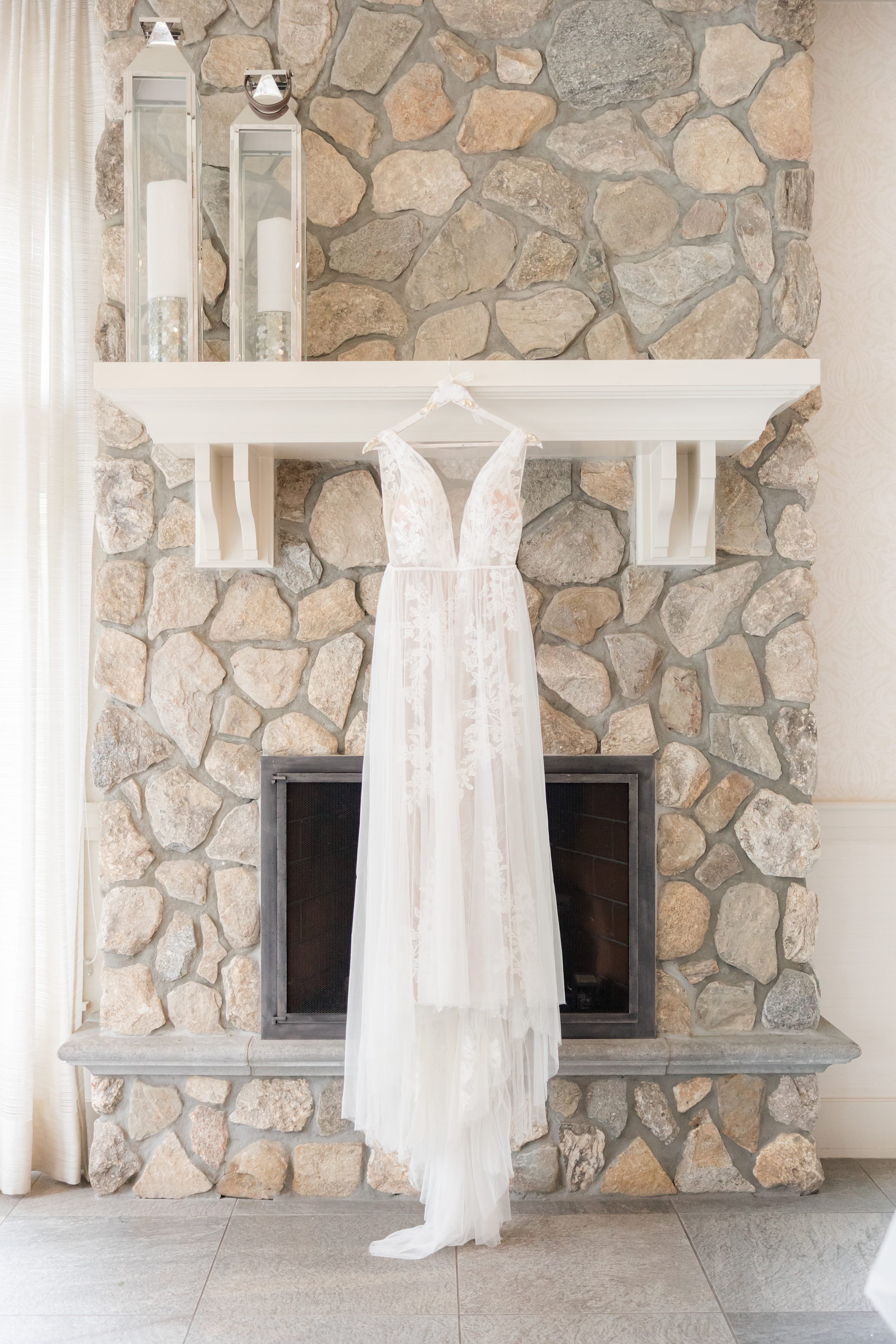 Romantic lace wedding dress hangs softly before a rustic stone fireplace, bathed in gentle light, evoking timeless elegance and the quiet anticipation of a bride’s special day