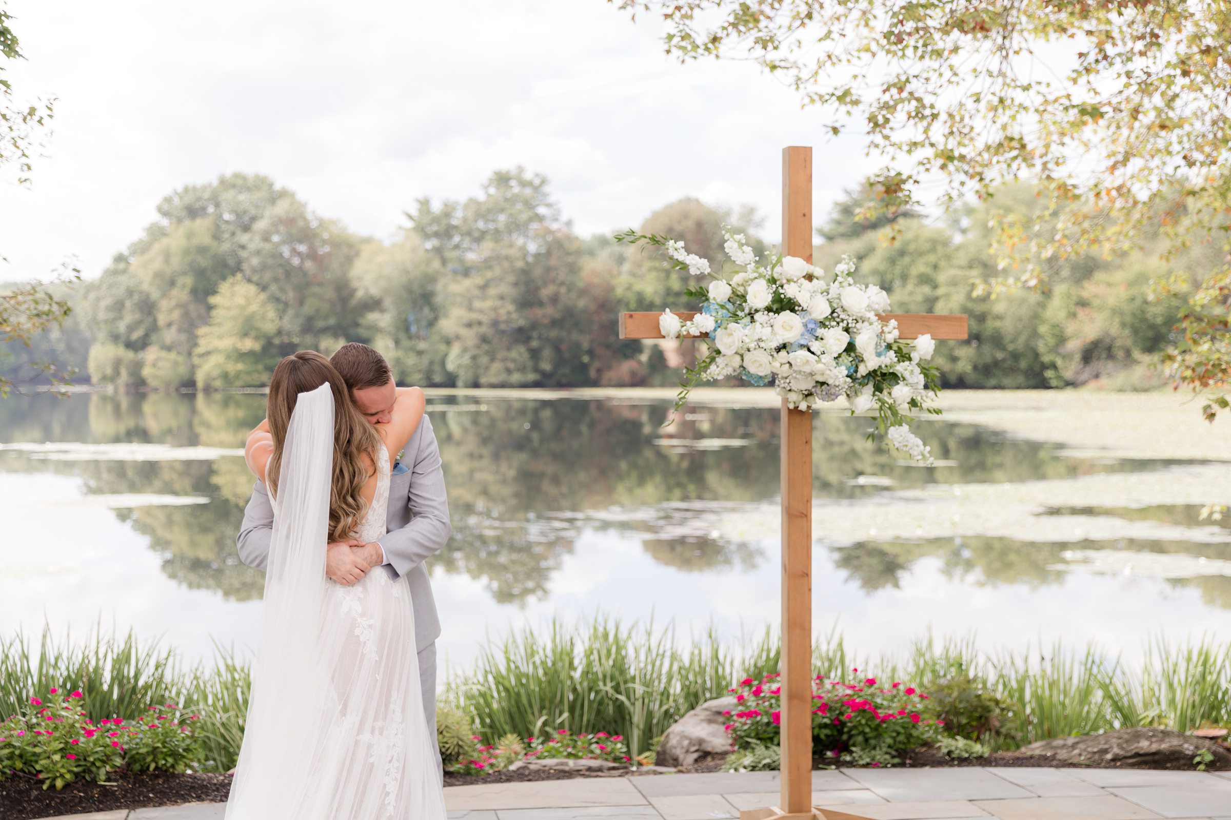 Beneath a flower-adorned cross by the still water, the bride and groom share a quiet kiss, wrapped in a sacred, tender moment of love and devotion