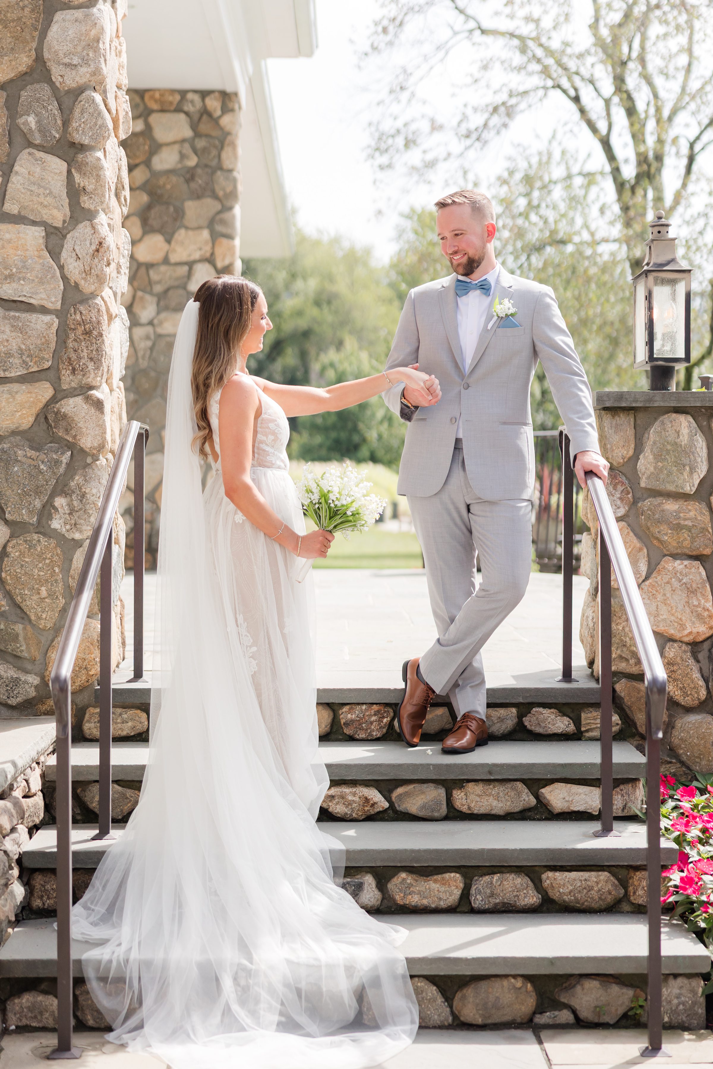 Bride reaches for her groom’s hand on sunlit stone steps, their eyes meeting in a tender, joy-filled moment as they step into forever together