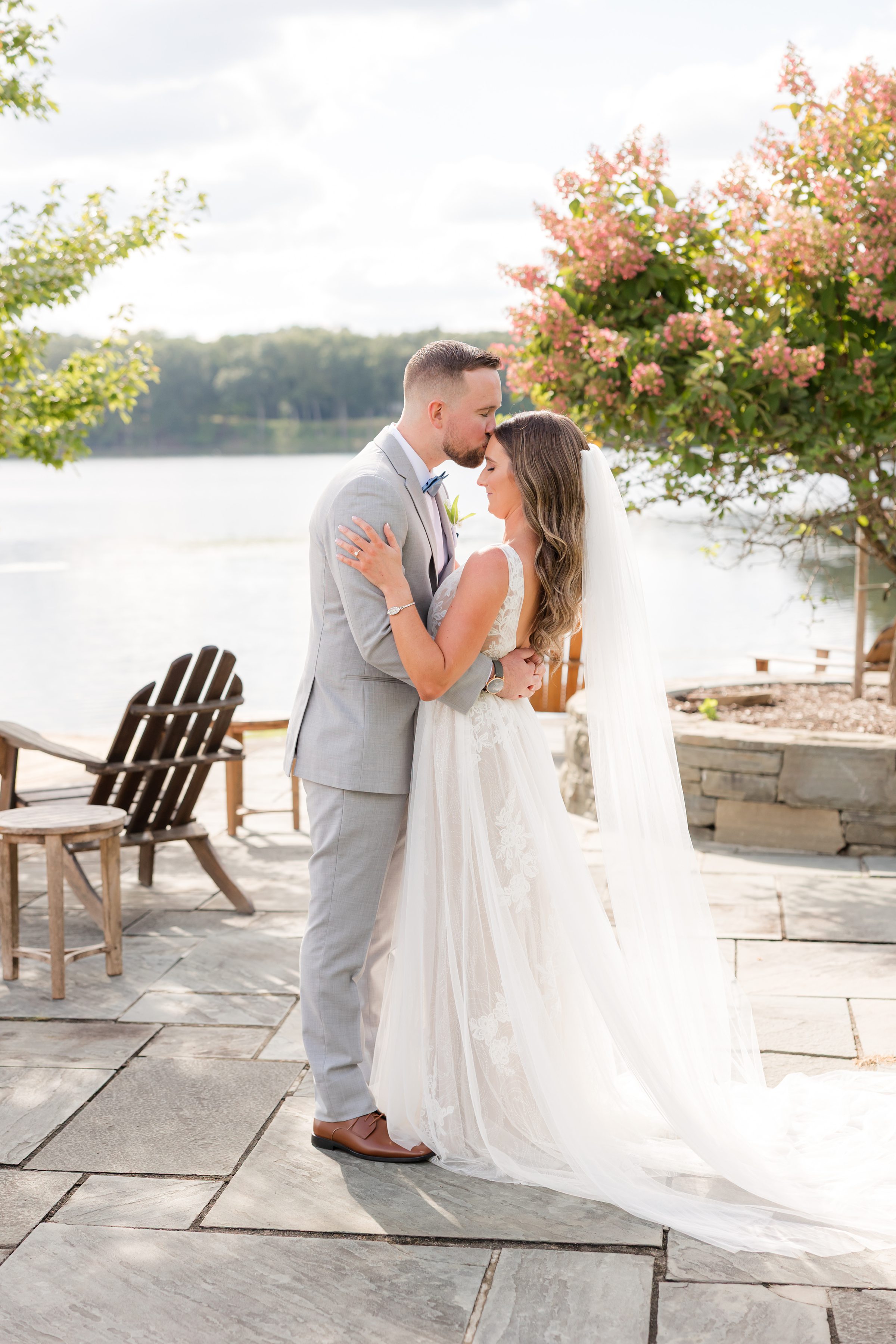 Bride and groom share a quiet, intimate embrace by the water, foreheads gently touching as soft light, blooming flowers, and still reflections surround their moment of love