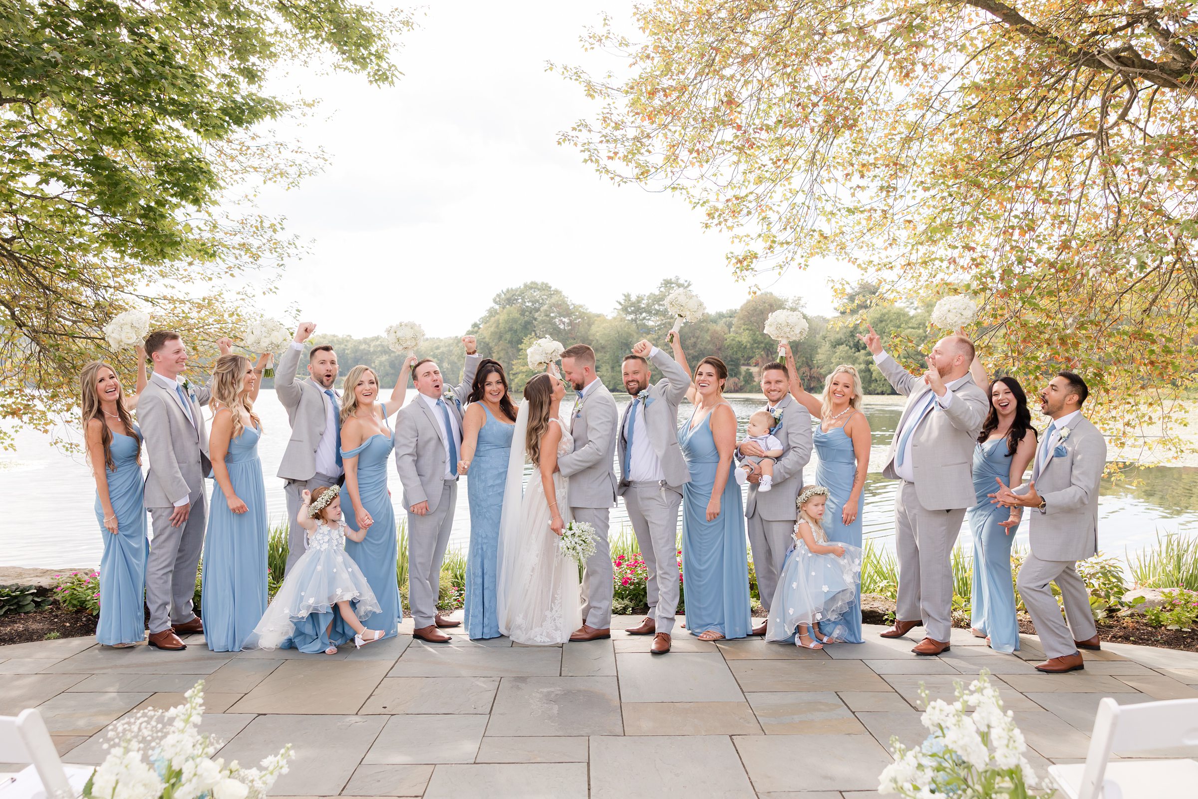 Bride and groom stand at the center of their wedding party, surrounded by loved ones in soft blue and gray tones, sharing a joyful moment beneath sunlit trees by the water