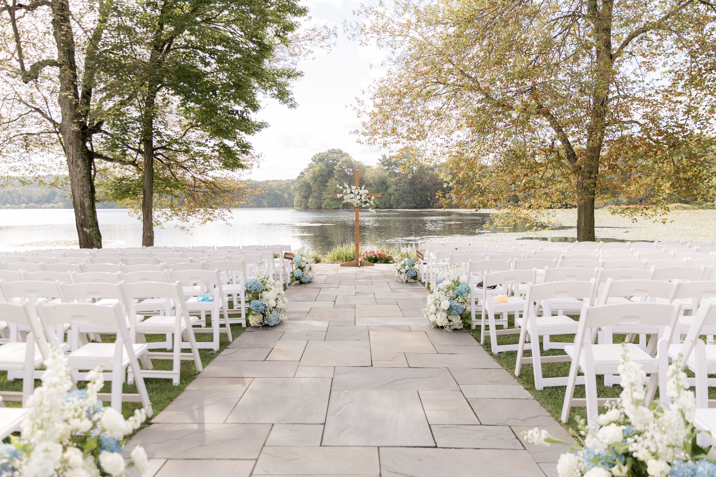 Serene aisle of white chairs and fresh florals leads toward a tranquil lake beneath glowing trees