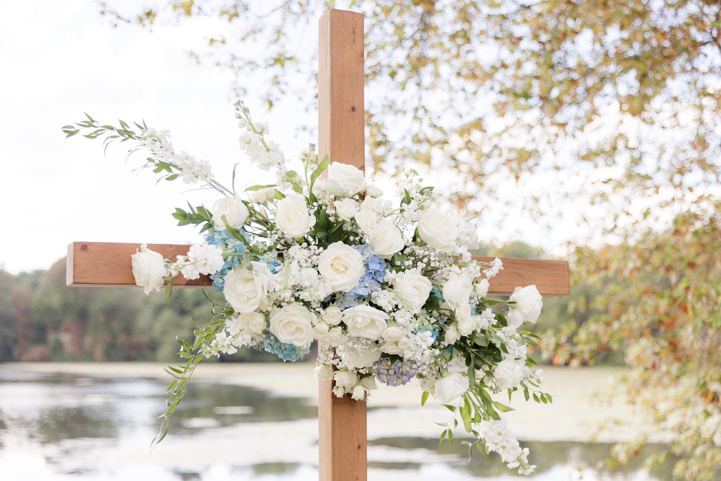 Wooden cross adorned with delicate white blooms overlooks the peaceful lakeside ceremony