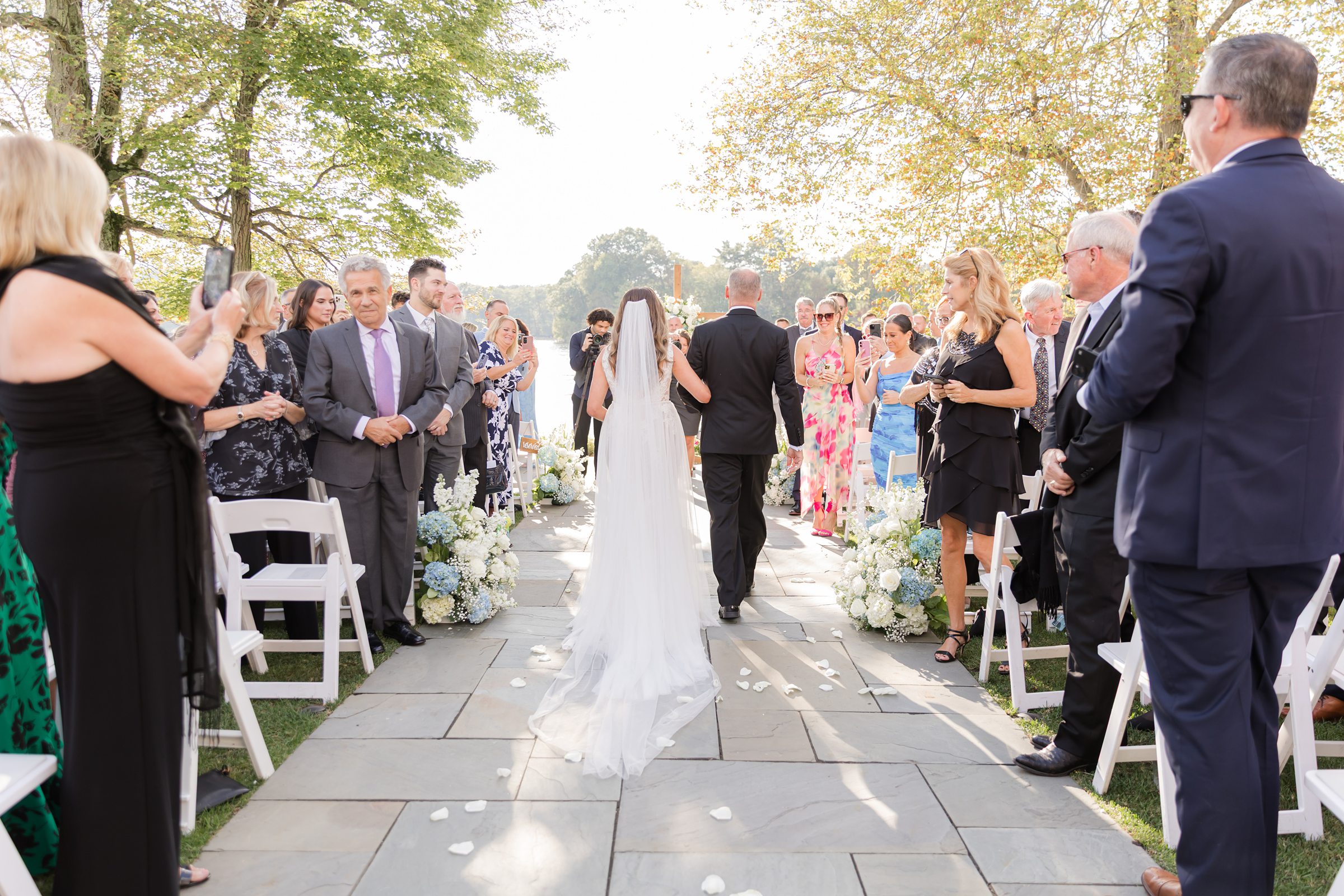 Bride walks down the aisle with her father, her gown flowing behind her as loved ones look on, surrounded by soft sunlight and blooming florals