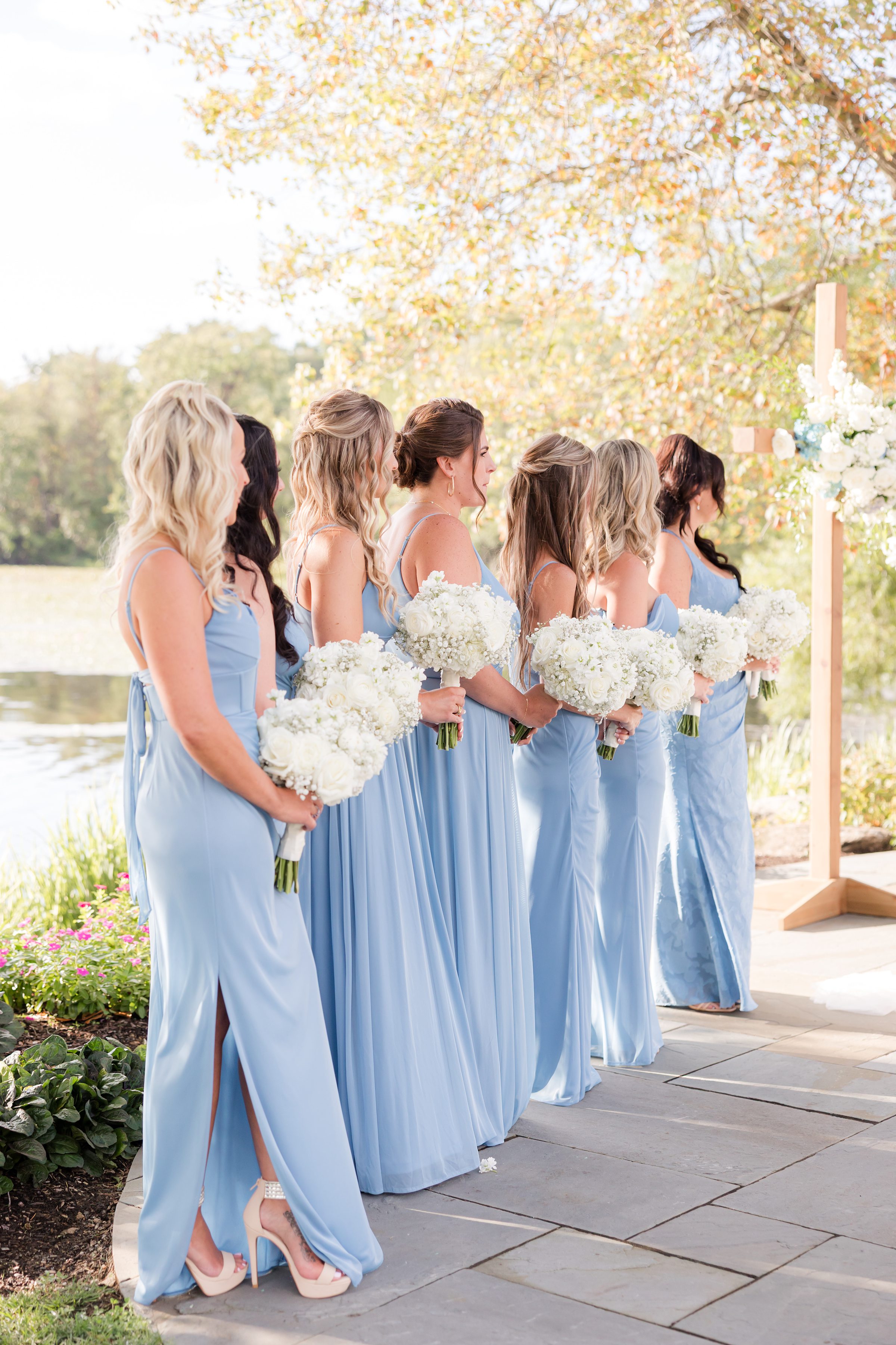 Bridesmaids in flowing blue dresses hold white bouquets, standing gracefully as they watch the ceremony by the water