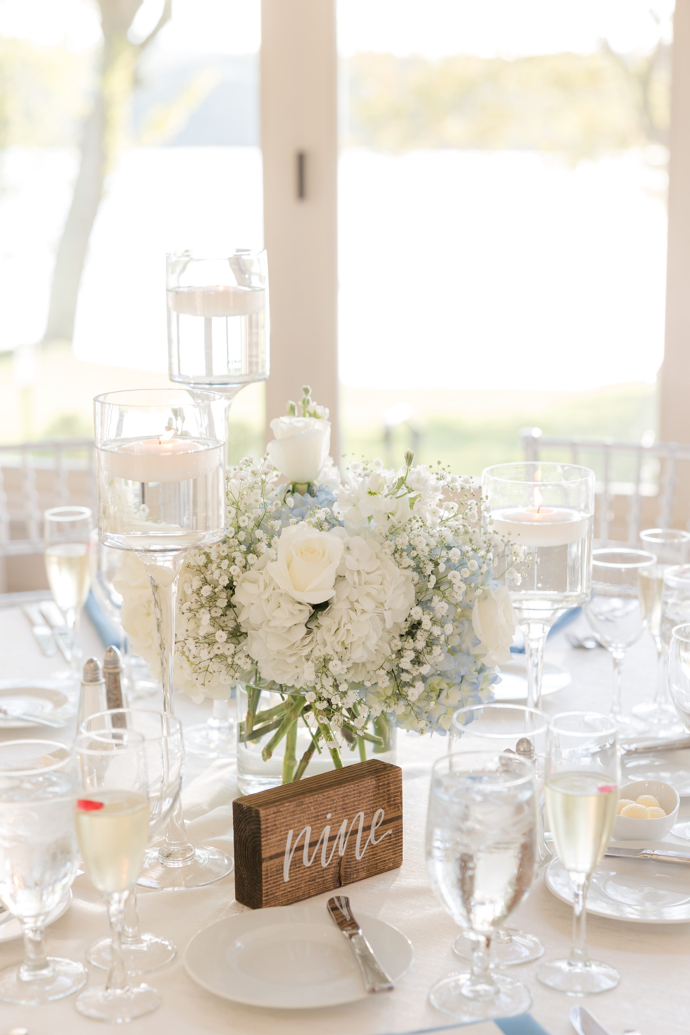 Romantic, softly lit reception table with white floral centerpiece, glassware, and candles, featuring a “Mr & Mrs” sign by a bright window overlooking the water