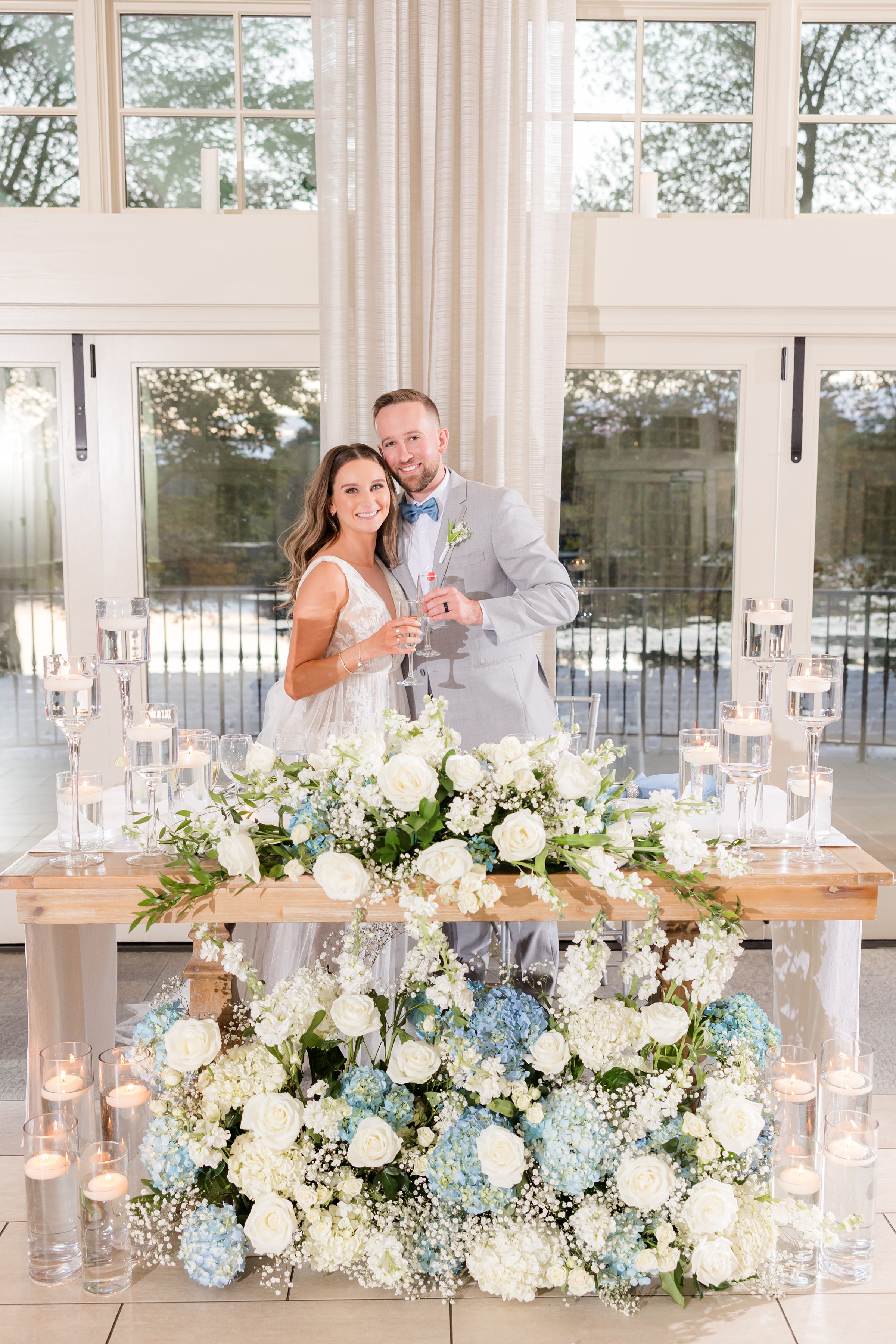 Newlyweds standing behind a decorated sweetheart table with white and blue floral arrangements and candles