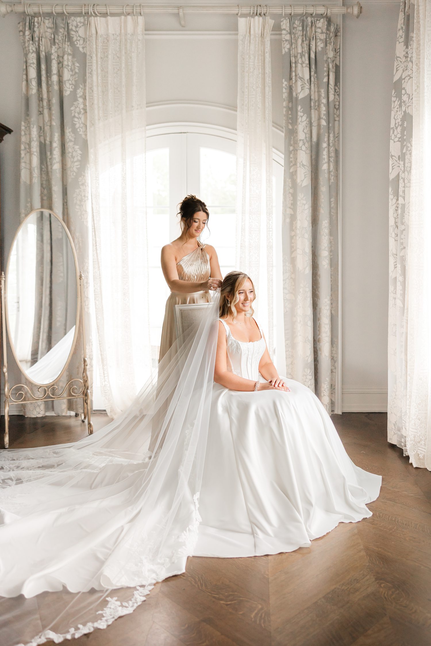 bride in elegant, voluminous white gown with her maid of honor fixing her veil near a large window draped with soft, sheer curtains