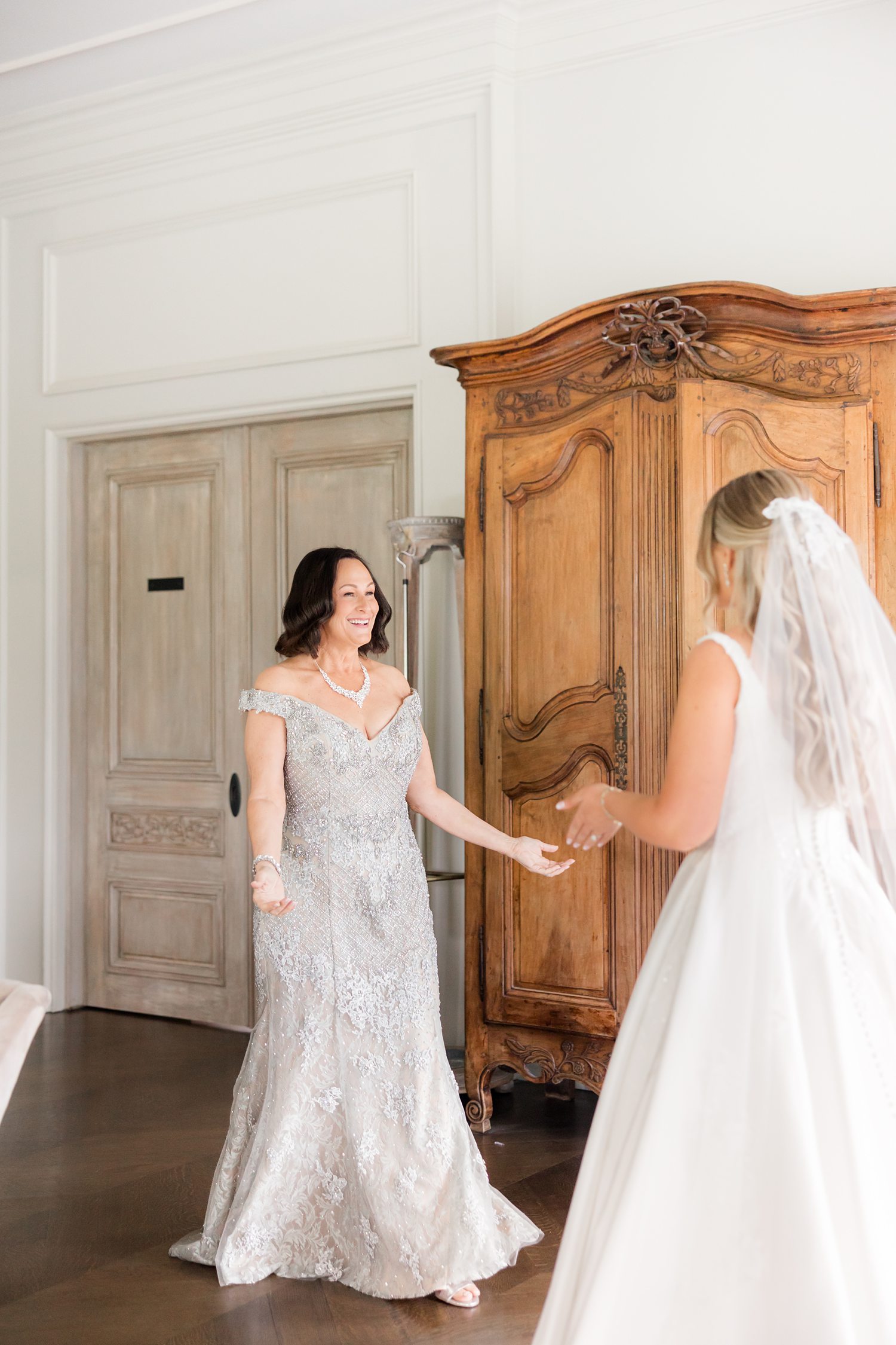A back view of the bride in her elegant, white sleeveless wedding gown and veil, captured as she welcomes her mother into the bridal suite.