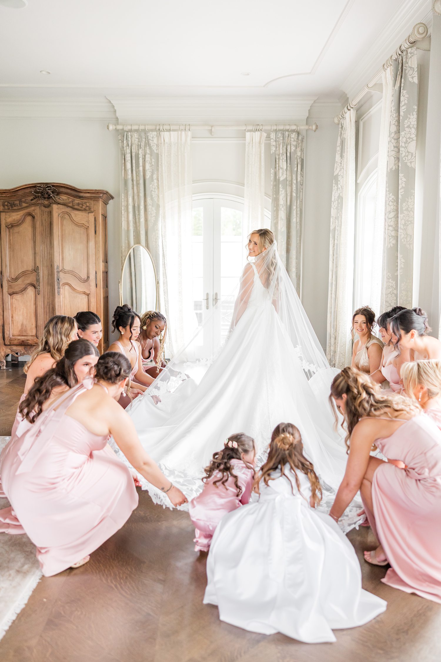 bride is wearing a stunning white sleeveless ballgown paired with an elegant veil that complements her sophisticated bridal look surrounded by her bridesmaids and flower girls, creating a joyful and cohesive wedding scene arranging her veil