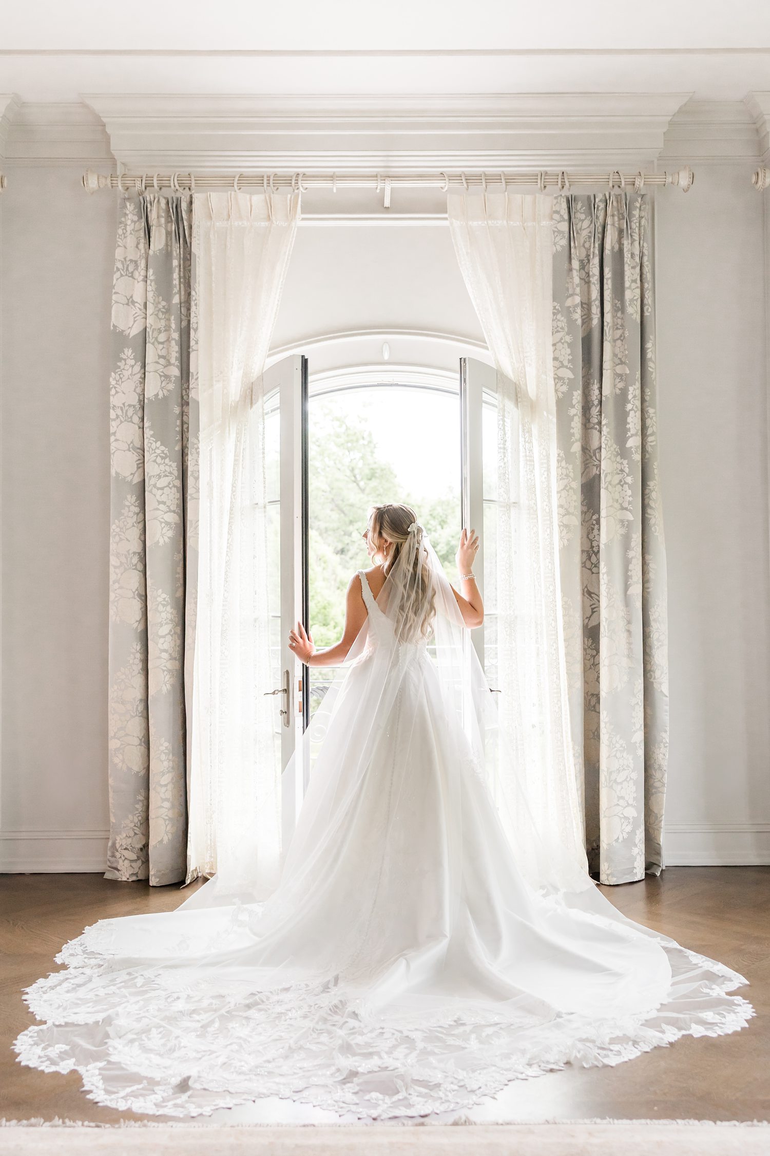 Back view of the bride posing as she opens the large white windows, capturing an angelic and elegant portrait that highlight her veil trained to the floor