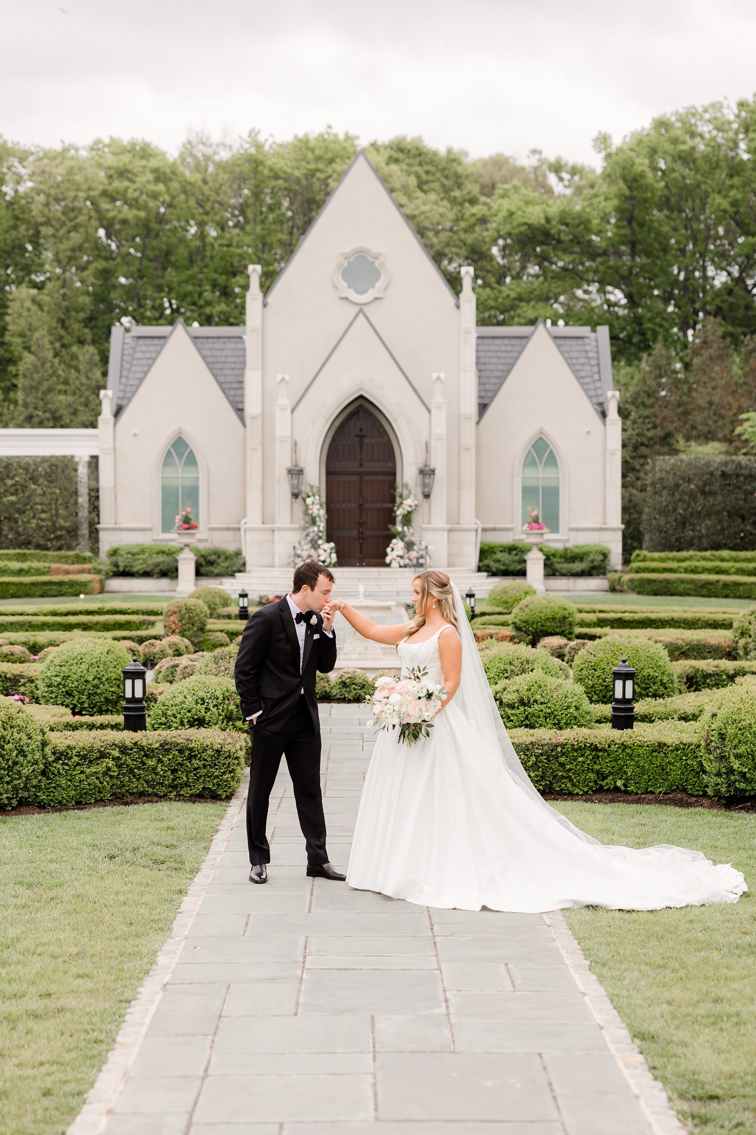 Bride and groom standing along a stone path in a manicured hedge garden, with the groom gently kissing the bride’s hand in front of the white Park Chateau Estate chapel
