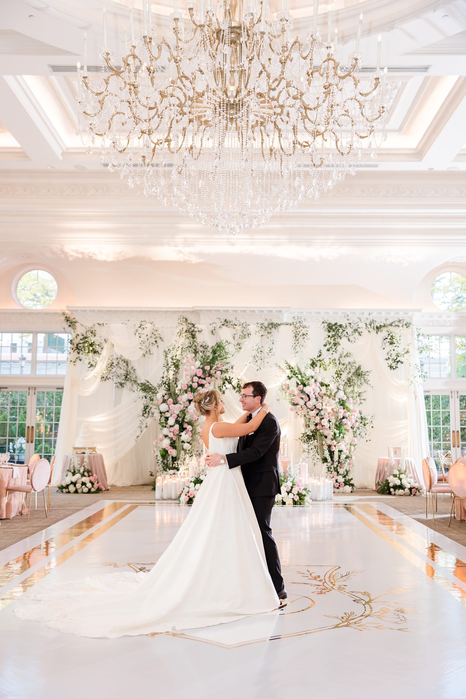 bride and groom share a romantic first dance under an elegant crystal chandelier, centered on a polished white dance floor, with a subtle gold monogram detail