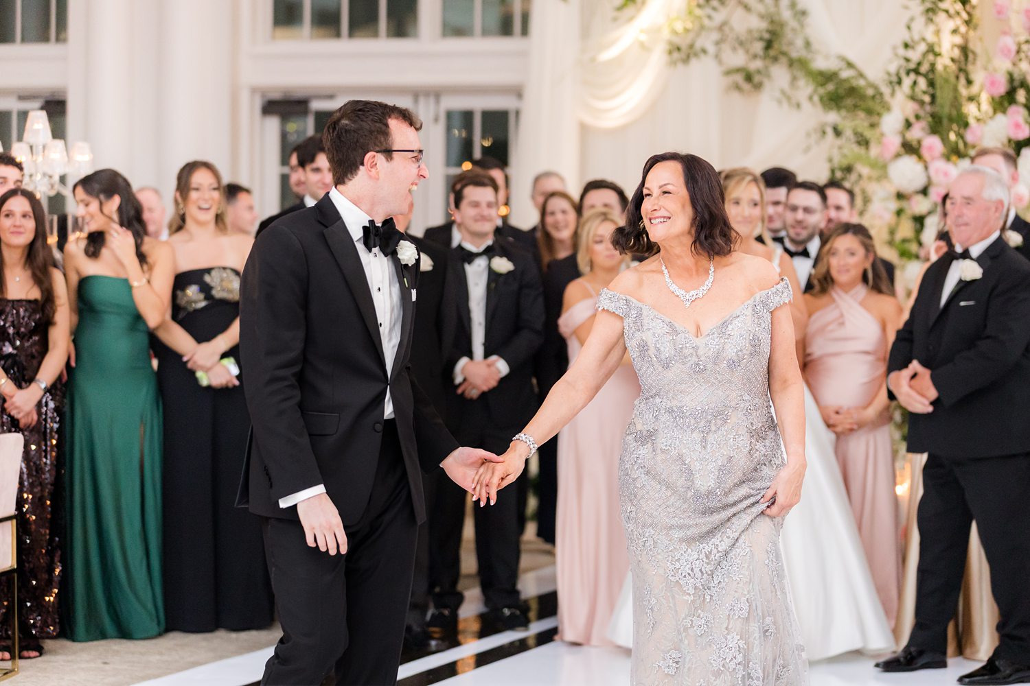 Wedding moment of groom sharing a dance with his mother admired by guests surrounding the ballroom reception