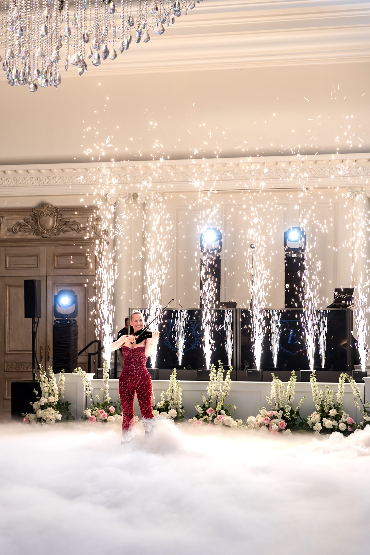 Wedding entertainment showcasing a violin performer amidst a thick layer of low-lying white fog Five tall with vertical jets of white-gold sparklers erupt from the stage behind