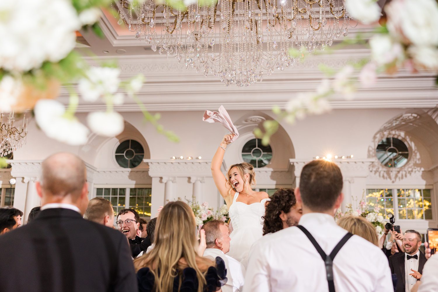 Inside an elegant, brightly lit venue with crystal chandeliers, a bride raises her pink cloth in celebration surrounded by guests capturing the moment on their phones
