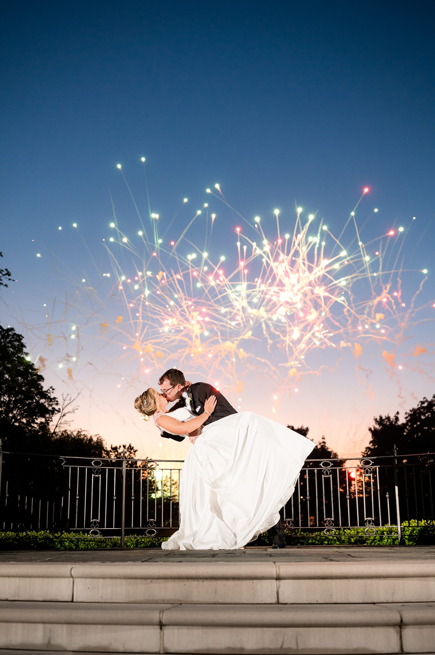 vertical photograph of a newlywed couple kissing passionately on an outdoor stone patio at dusk, backdropped by a dramatic display of colorful fireworks illuminating the evening sky