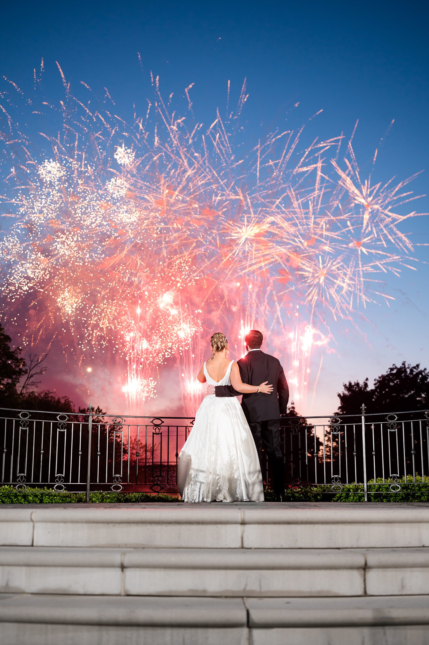 bride and groom stands on stone steps, looking away toward a vibrant display of red, orange, and blue fireworks illuminating the night sky above a dark tree line