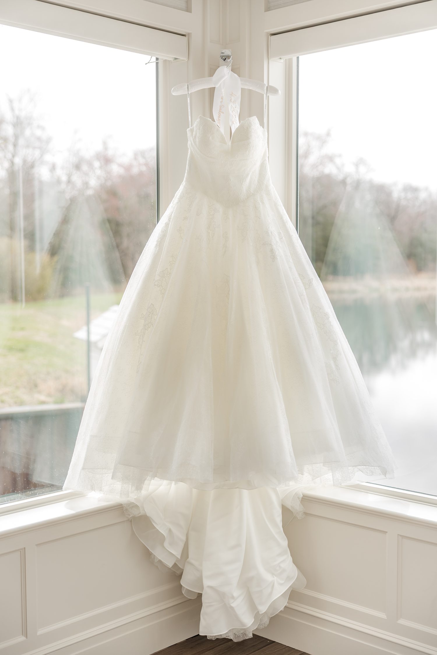 Elegant white wedding dress hanging by large window in lakeside manor bridal suite