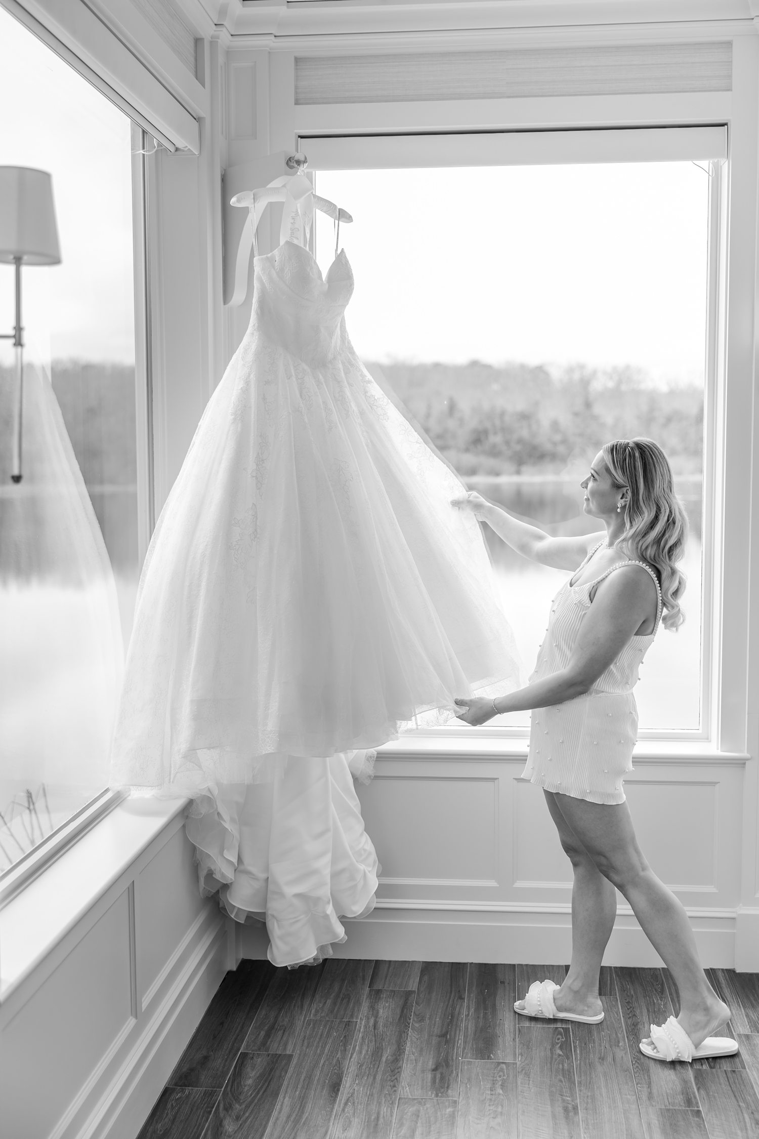 Black and white photo of bride adjusting wedding dress by bright window