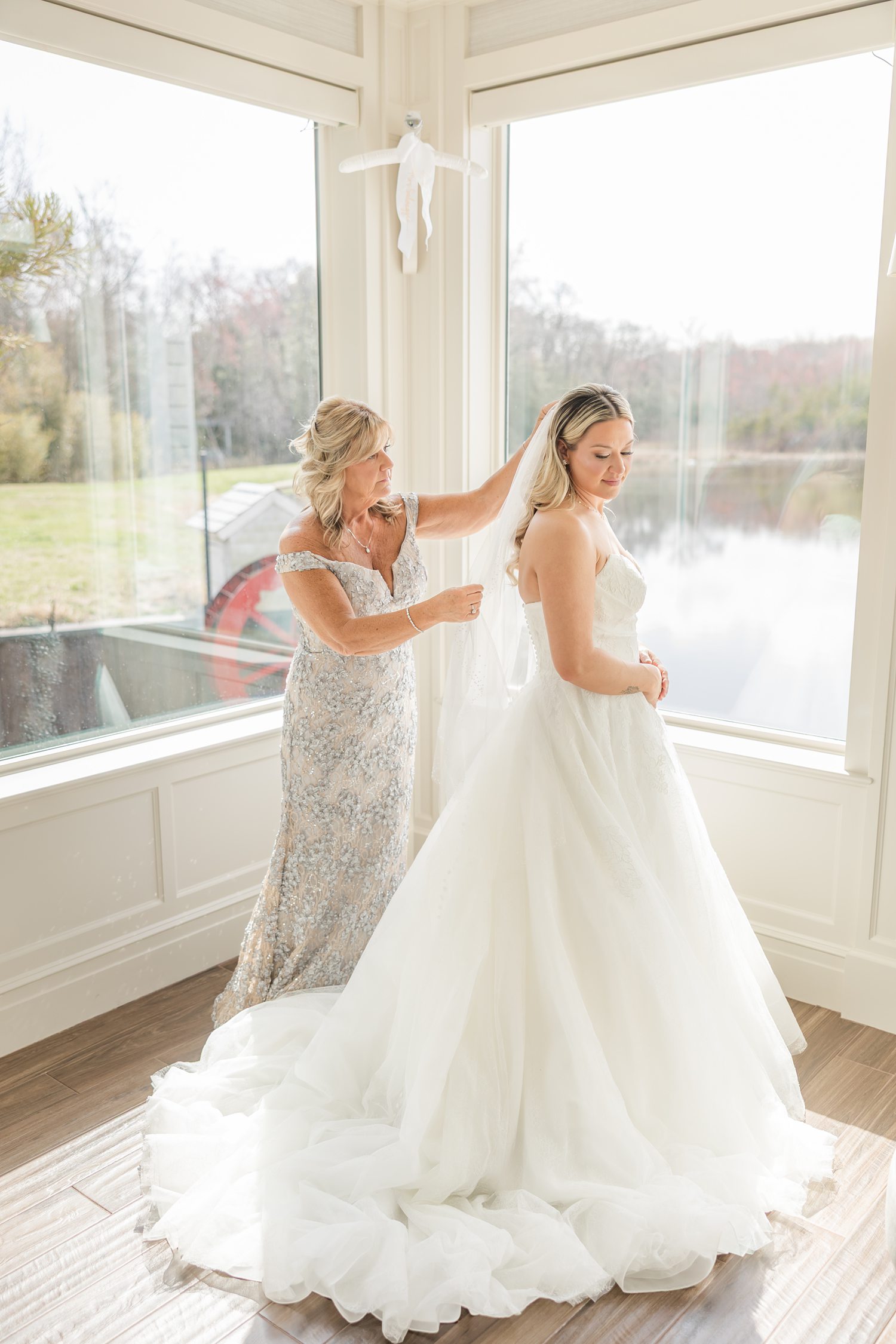 Bride and bridesmaid adjusting wedding gown near window before ceremony