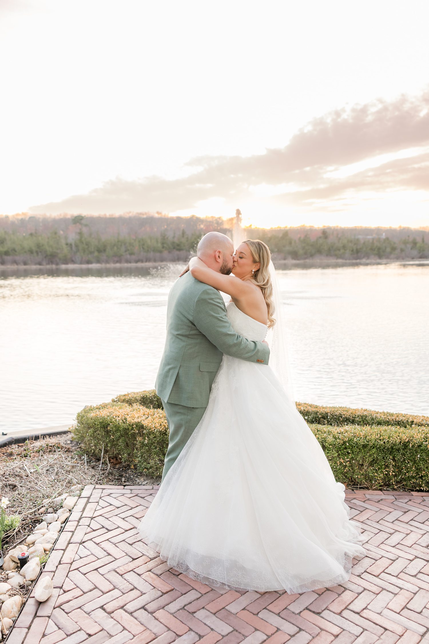 Newlywed couple embracing and kissing by the water during romantic sunset wedding portraits
