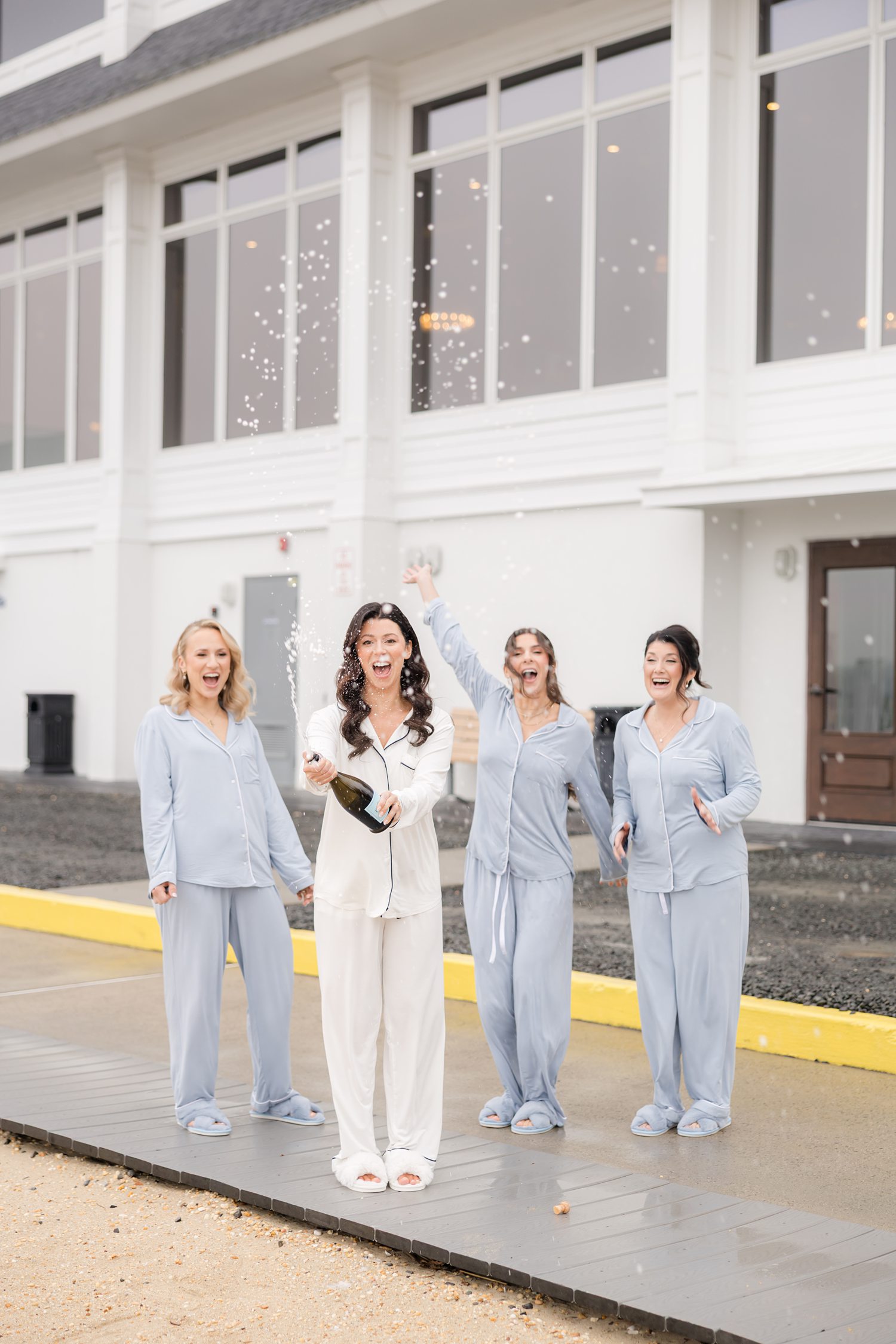 Bridesmaids in light blue pajamas outside, cheering while the bride pop a champagne 