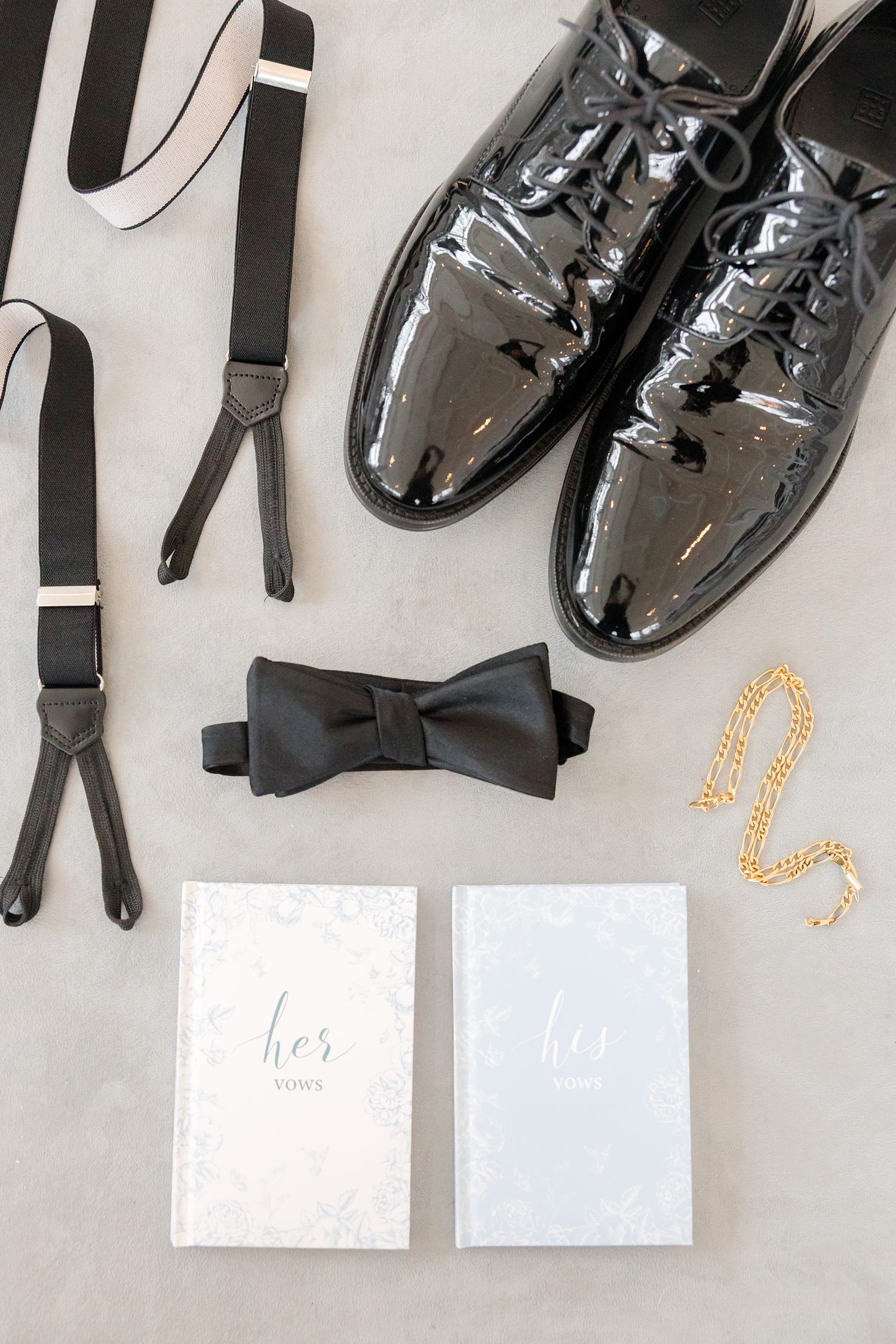 Groom’s details including black shoes, bow tie, suspenders, gold bracelet, and wedding vow books