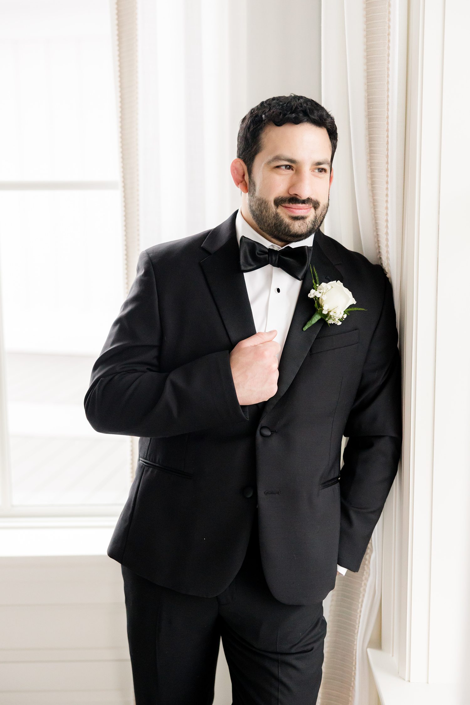 Groom's portrait in black tuxedo leaning against the window