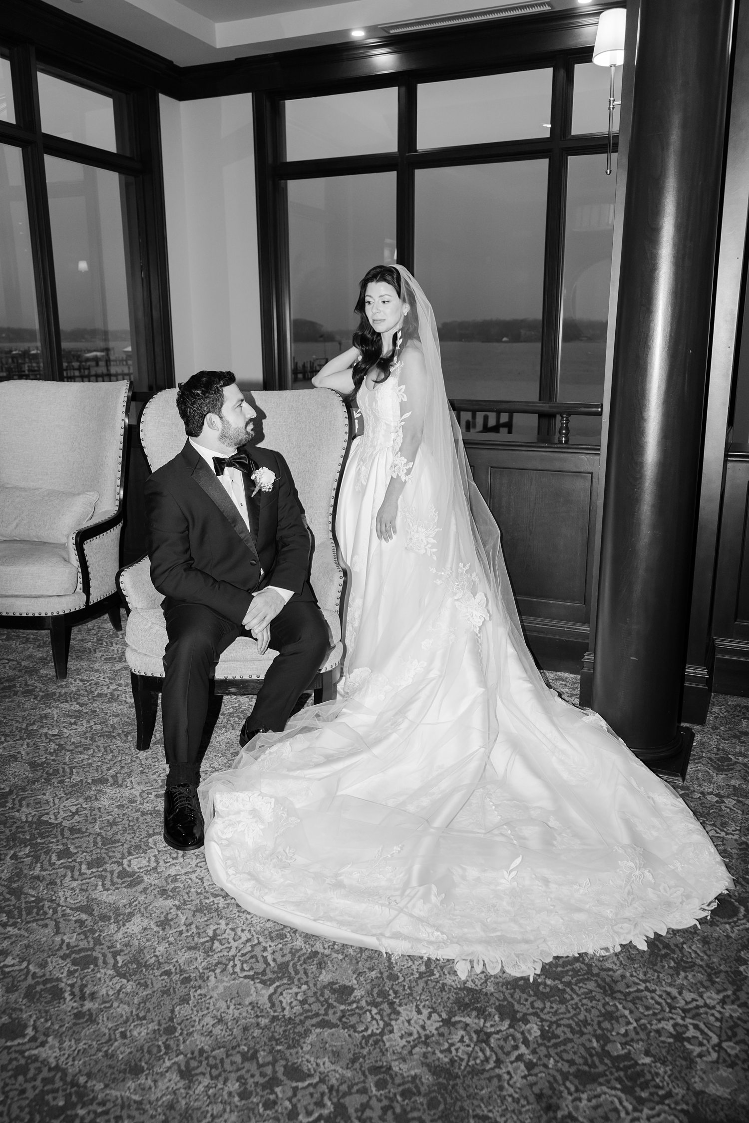 Black and white portrait of the groom sitting while the bride is standing leaning on the chair in the lounge