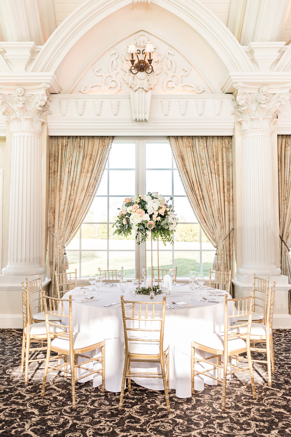 Round reception table with tall floral centerpiece beside bright windows at Ashford Estate